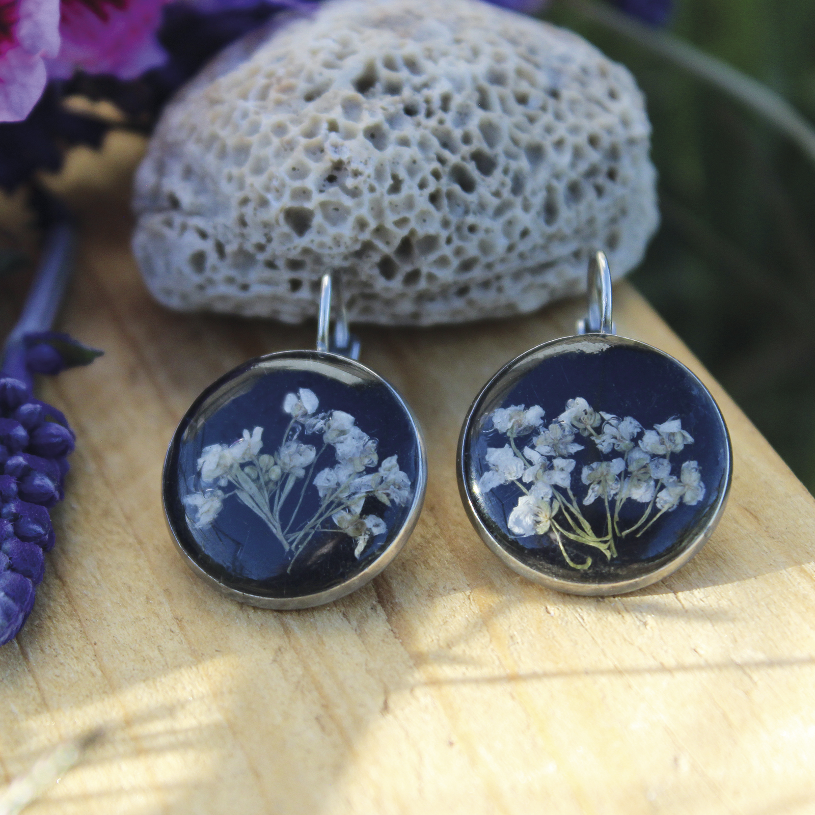 Pendientes redondos grandes de acero hipoalergénico con flores de aliso marítimo naturales en resina sobre fondo negro, hechos a mano en Malas Herbas Artesanía, artesanos gallegos.