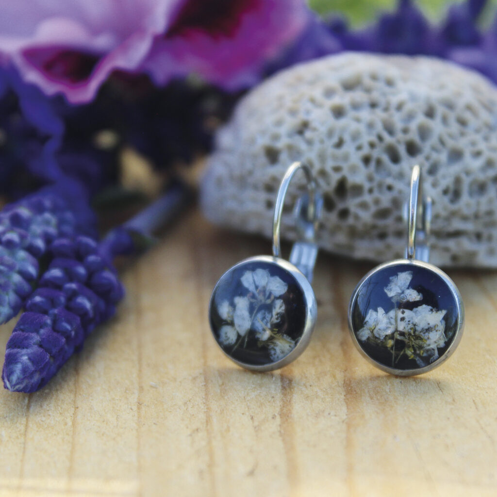 Pendientes redondos pequeños de acero hipoalergénico con flores de aliso marítimo naturales en resina sobre fondo negro, hechos a mano en Malas Herbas Artesanía, artesanos gallegos.