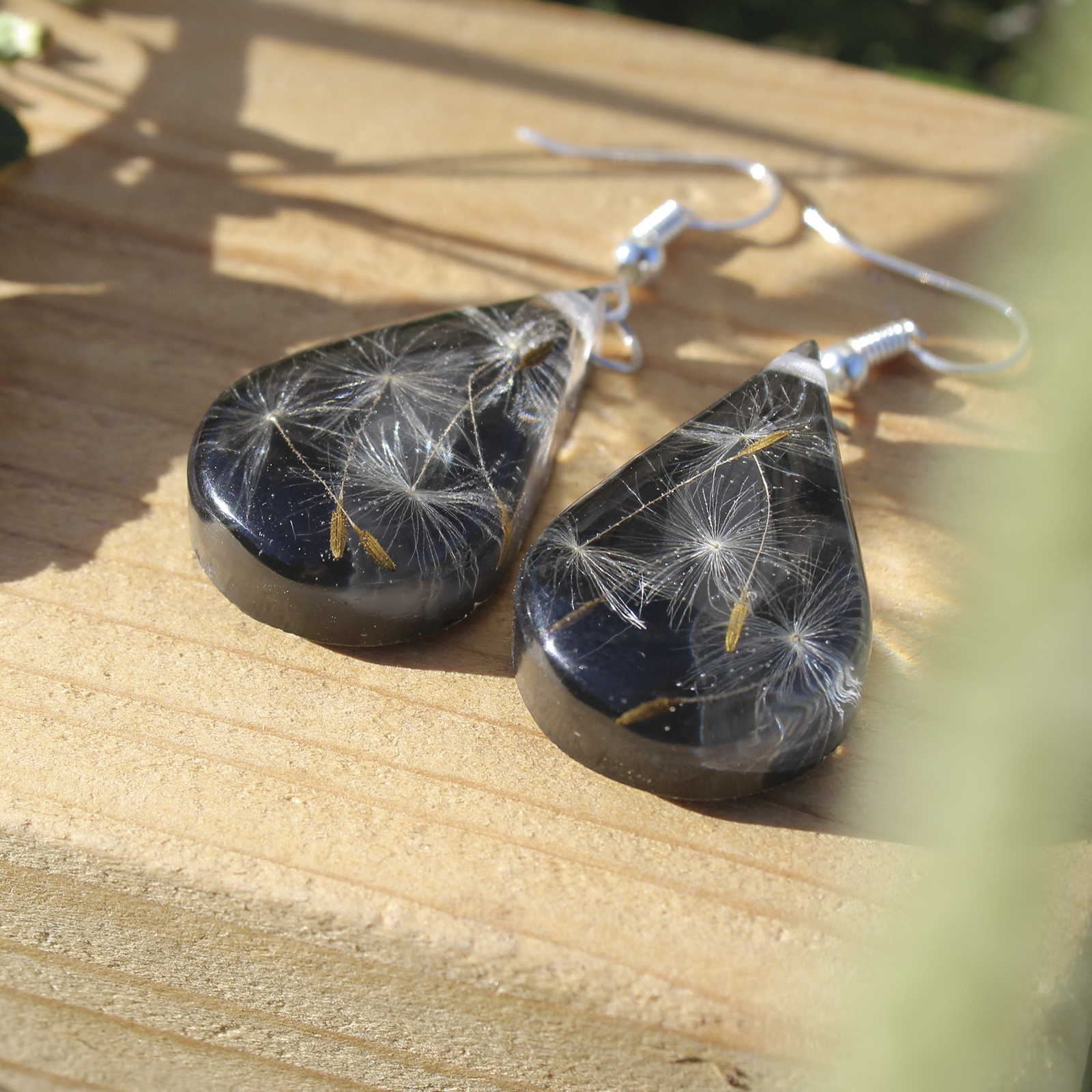 Pendientes de gota en resina con semillas de diente de león naturales sobre fondo negro, hechos a mano en Malas Herbas Artesanía, artesanos gallegos.